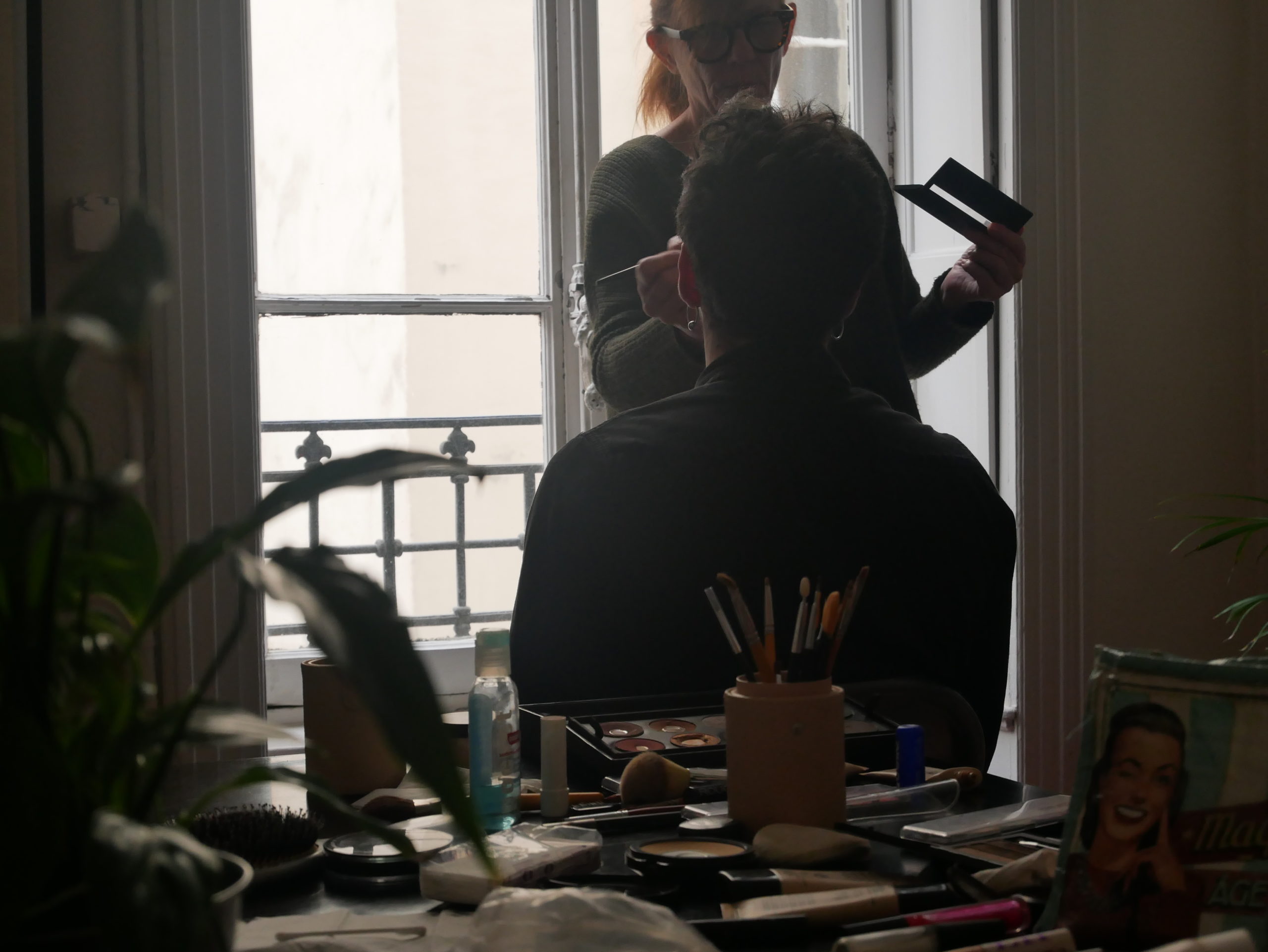 Séance de maquillage pour le shooting photo de l'agence avec accessoire en premier plan étalé sur une table et personne de dos se faisant maquiller