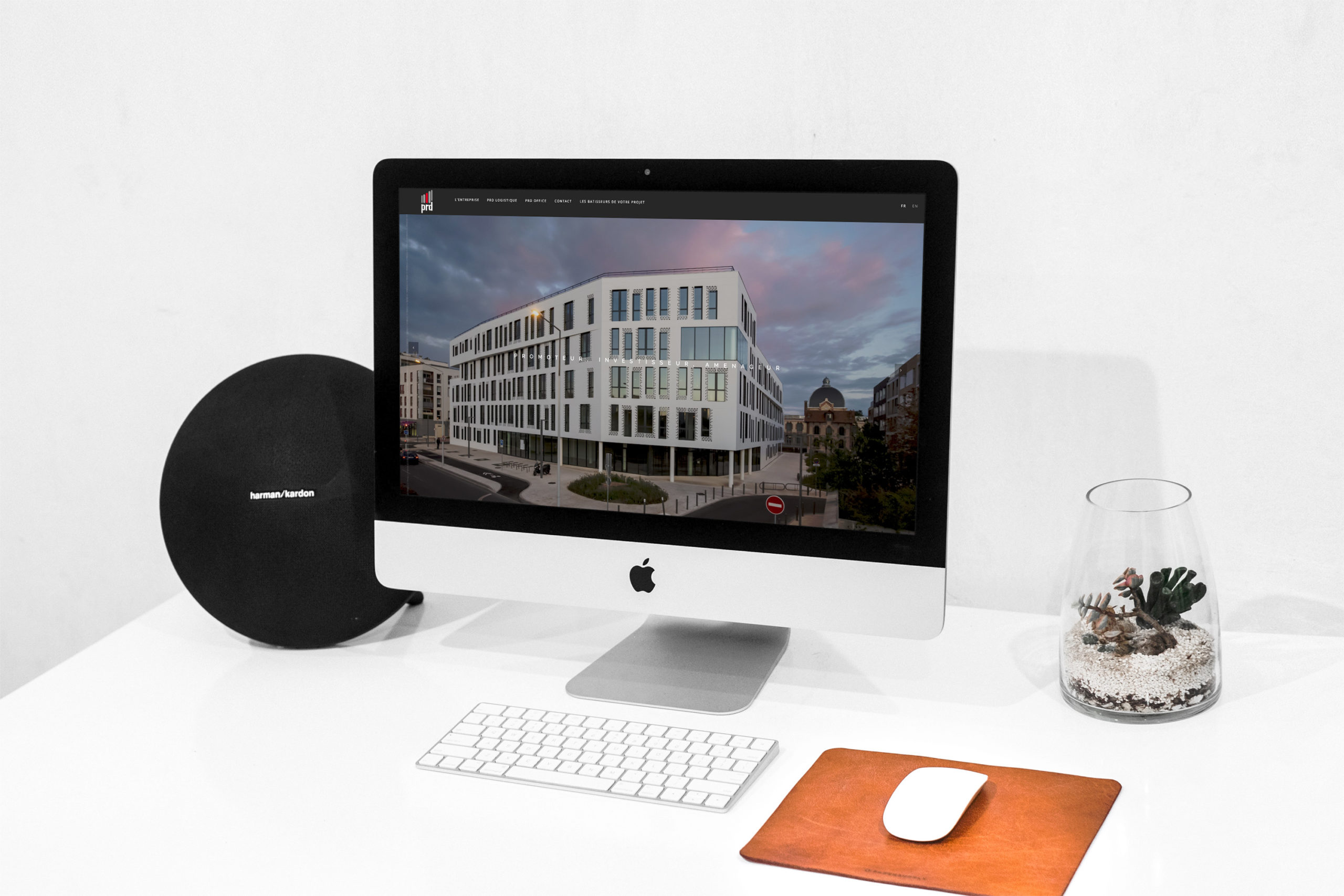 Mise en situation du site internet de PRD sur un ordinateur fixe de type MAC avec une enceinte et une plante verte à côté