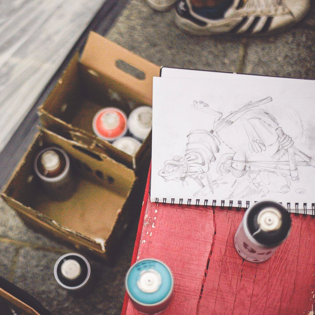 Gros plans sur le sol avec des basket adidas, des bombes de peintures de plusieurs couleurs dans une caisse et un dessin au crayon de papier qui va devenir une fresque