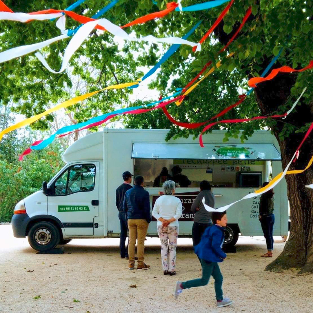 Personnes qui attendent devant un food truck pour commander à mange sous un arbres avec des rubans de toutes les couleurs au vent